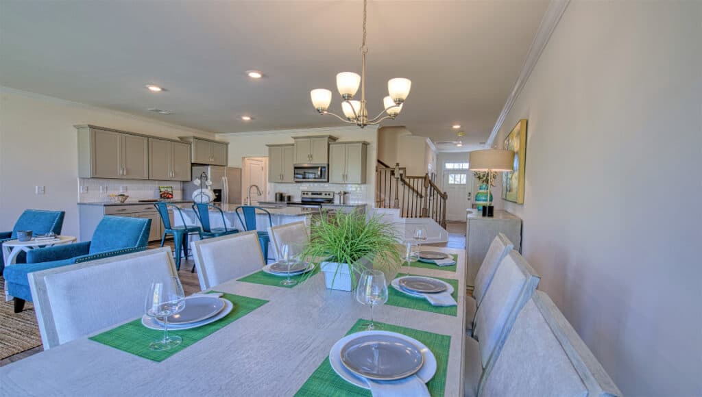 Elegant dining area featuring a long wooden table set with white plates, green placemats, and glasses, surrounded by blue upholstered chairs. A chandelier hangs above. In the background is a modern kitchen in The Townes at Stonecrest, an ideal Asheville retreat for senior living or retirement bliss.