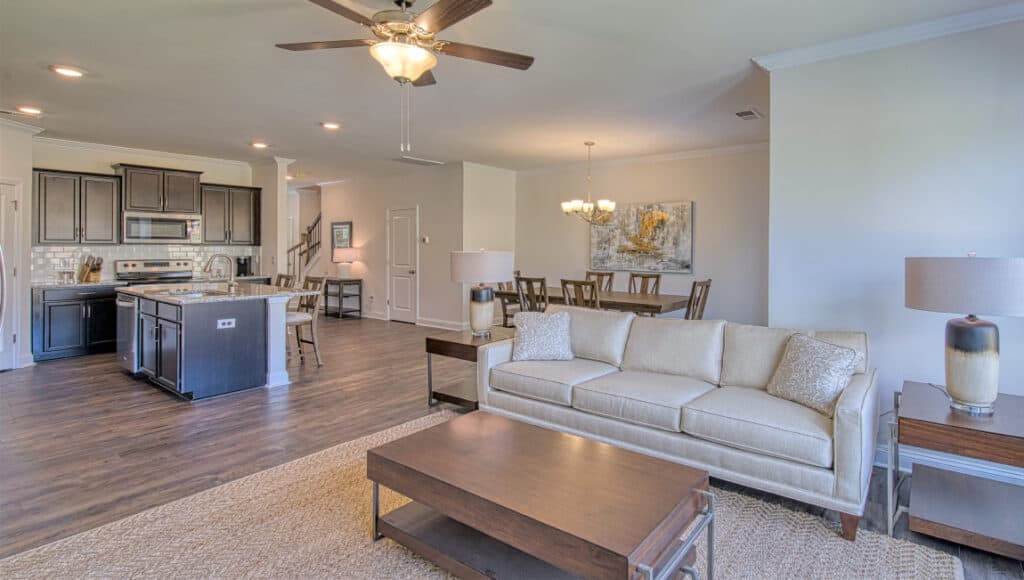 The Townes at Stonecrest offers an open-concept living room and kitchen, perfect for senior living. It features a beige couch, wooden coffee table, dining table, dark wood cabinets in the kitchen with an island bar seating. Ceiling fans and lamps light up the space while hardwood floors ensure easy maintenance.