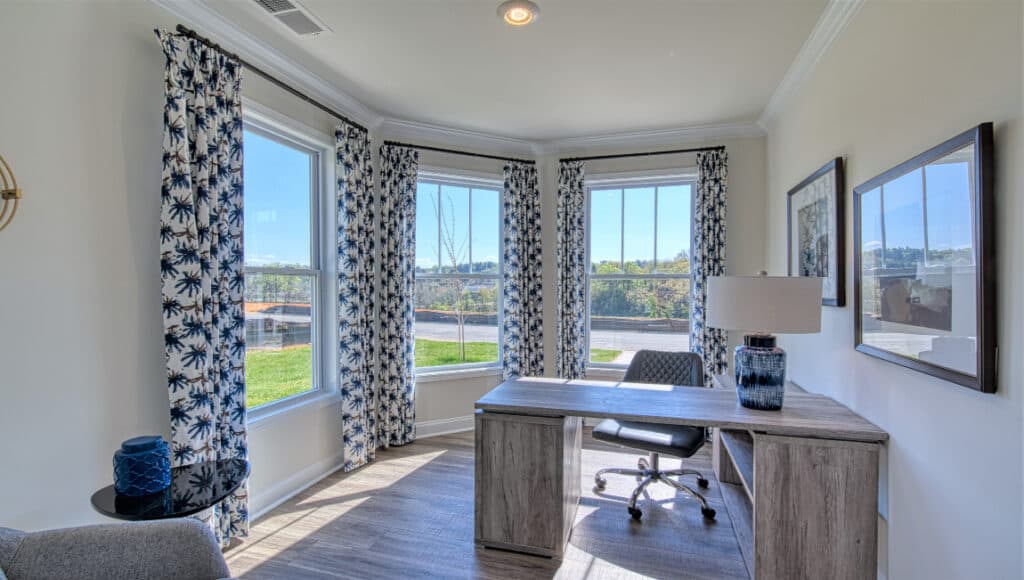 The Townes at Stonecrest features a bright home office with large windows and blue-patterned curtains. It includes a wooden desk, lamp, black office chair, and framed wall art. Sunlight illuminates the wooden floor, creating an ideal space for independent living pursuits in Hendersonville.