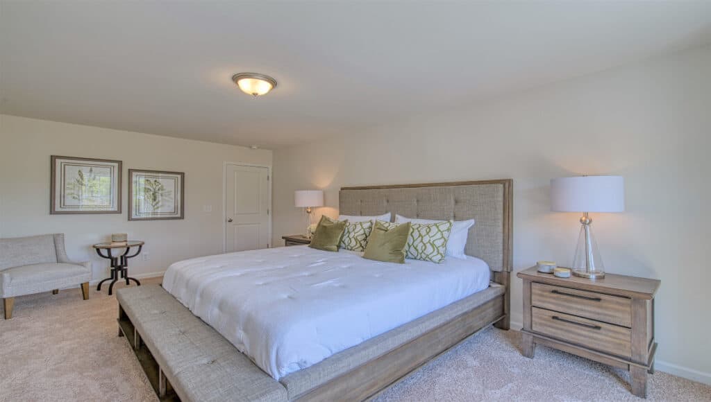 The Townes at Stonecrest offers a bright senior living bedroom with a large bed featuring a padded headboard. Two wooden side tables with lamps flank the bed. A cozy seating area in the corner has two chairs and a table, complemented by wall artwork, all in neutral tones.