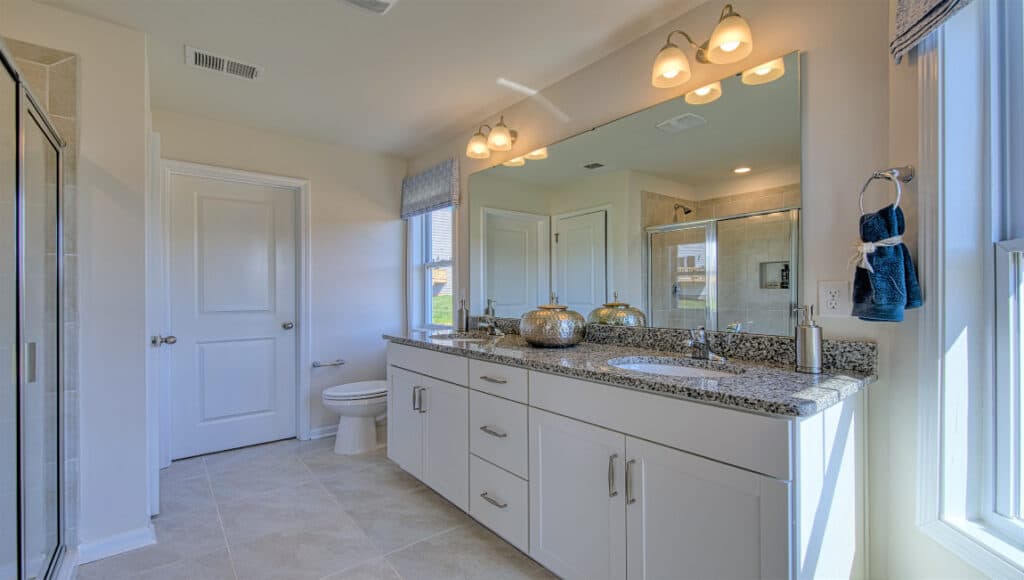 The Townes at Stonecrest in Hendersonville offers a modern bathroom ideal for independent living, featuring a large mirror above a double sink on granite, white cabinets for storage, and ample light from ceiling lights and windows—perfect for comfortable retirement living.
