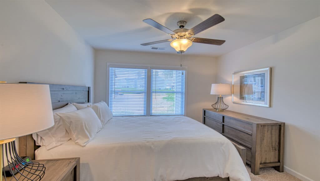 The Townes at Stonecrest in Hendersonville boasts a bright bedroom with white-linen bed, wooden nightstands and lamps, matching dresser with framed picture above, large window with blinds, and ceiling fan/light fixture—ideal for independent living.