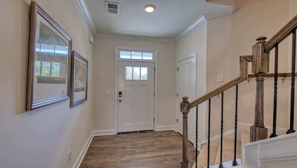 The Townes at Stonecrest features a wooden-floored hallway leading to a white front door with an overhead window, perfect for independent living. Two framed pictures enhance charm, while a staircase with wooden railings and natural light from a side window evoke serene Asheville homes.