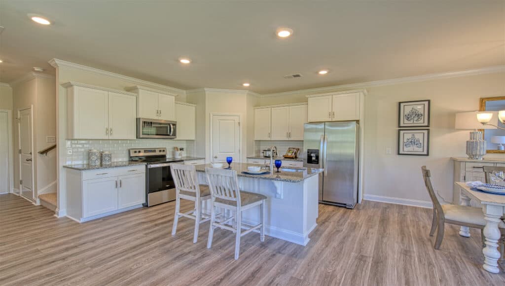 The Townes at Stonecrest offers a modern kitchen with white cabinets, stainless appliances, and a central island with two chairs. Ideal for independent living in Hendersonville, it connects to a dining area with a table, four chairs, and framed artwork. Light wood flooring and recessed lighting enhance the ambiance.