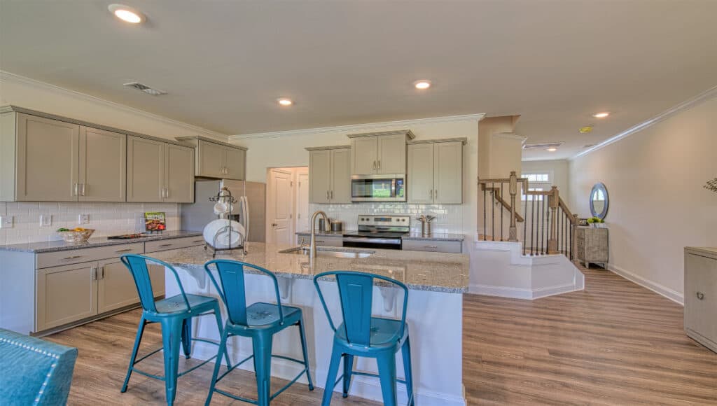 The Townes at Stonecrest features a sleek kitchen with granite countertops, white cabinets, and stainless steel appliances. Three blue metal stools line the central island. Wood flooring and recessed lighting enhance the warmth, ideal for independent living, with a staircase and hallway visible nearby.