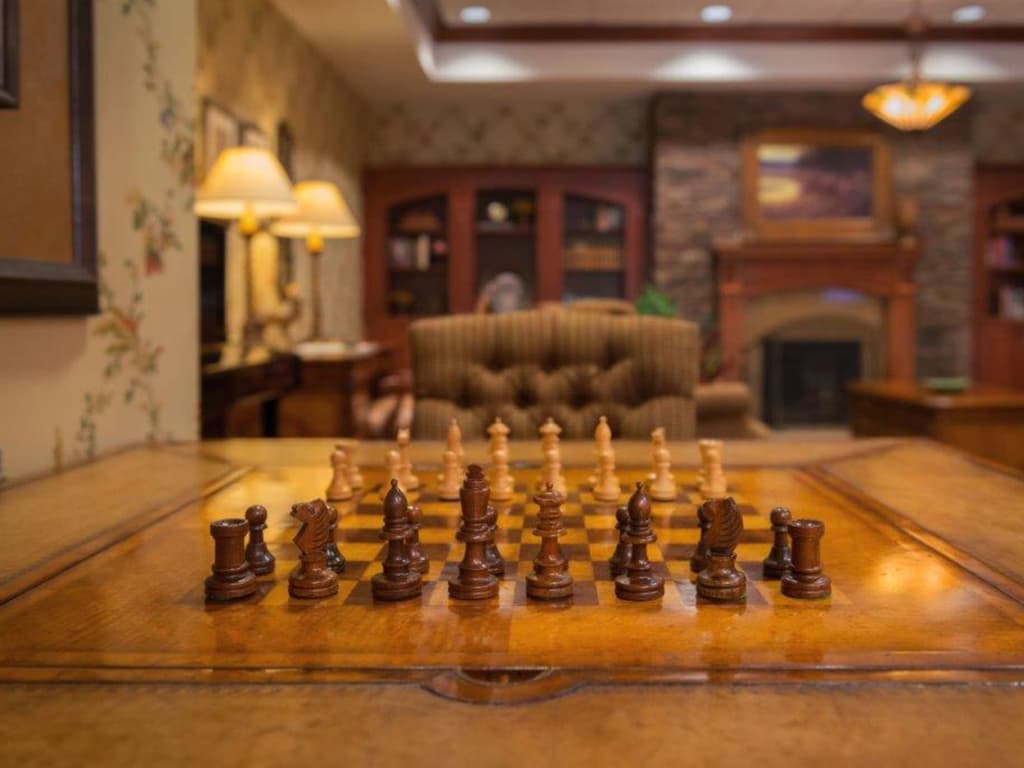 In a cozy senior living room at Lake Pointe Landing, a chessboard is set on a wooden table. Behind it, blurred bookshelves, a brown patterned armchair, and a fireplace create an inviting atmosphere reminiscent of Ashevilles charm.