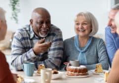 A group of older adults sit around a table, smiling and laughing together while enjoying coffee, cake, and pastries in a bright, cozy setting.
