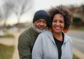 A smiling couple stands outdoors in Asheville. The man, wearing a black beanie and green sweater, embraces the woman from behind. She sports a gray jacket and orange earrings, perfectly capturing the joy of retirement life amidst the blurred street and trees in the background.
