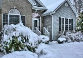 A snow-covered suburban house with brick and gray siding, perfect for retirement in Asheville. Large windows, a white door, and snow-laden bushes grace the front yard. Flakes gently fall as a path leads from the freshly shoveled steps, inviting cozy senior living amidst serene beauty.