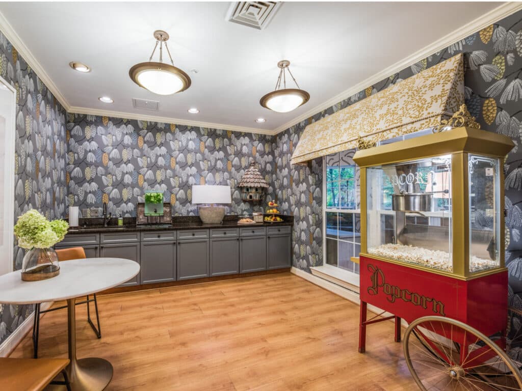 The cozy room at Arbor Terrace of Asheville boasts a popcorn machine, table, and chair. Leaf-patterned wallpaper and floral curtains adorn the space, enhanced by cabinets, a decor-adorned counter, and warm wooden floors—ideal for independent living.