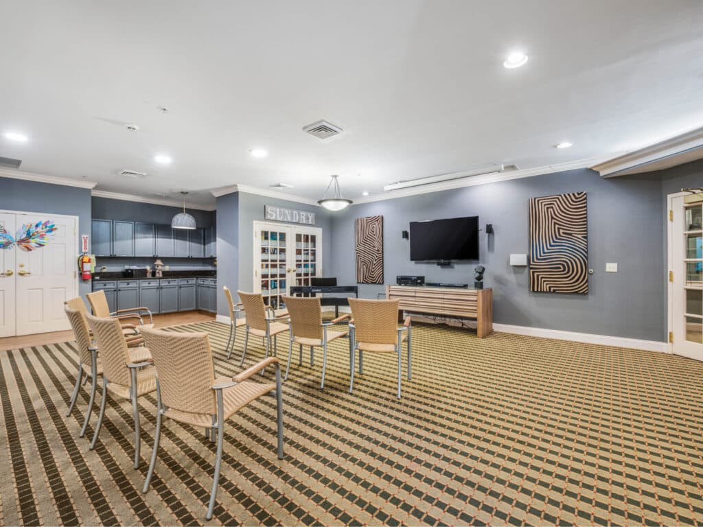 The Arbor Terrace of Asheville features a modern lounge with wicker chairs arranged towards a wall-mounted TV, patterned carpeting, abstract artwork, a corner kitchenette, and a door labeled Sundry. Recessed lighting enhances the cozy retirement atmosphere.