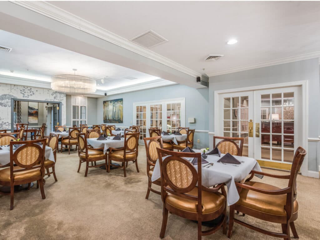 The Arbor Terrace of Asheville offers a spacious dining room with round tables, patterned wooden chairs, light gray tablecloths, and dark napkins. Light blue walls and plush carpet complement the rooms large circular chandelier—ideal for hosting a memorable retirement celebration.