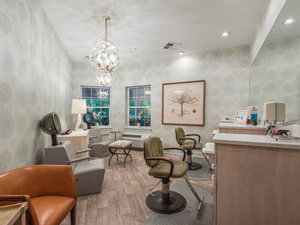Arbor Terrace of Asheville features a well-lit salon with modern decor, two stylist chairs, and hairdressing equipment. It boasts light walls, hardwood floors, a large mirror, elegant framed artwork, hanging lights, and windows showcasing lush greenery—perfect for a senior living community.