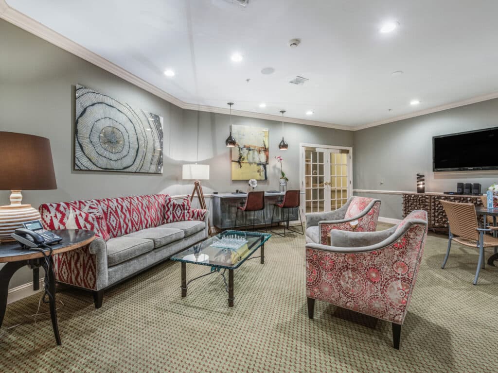 The Arbor Terrace of Ashevilles cozy living room features a red and gray patterned sofa, two red armchairs, and a glass coffee table. A large abstract painting adorns the wall, with a TV and wooden desk in the background; lamps provide warm lighting for restful retirement evenings.