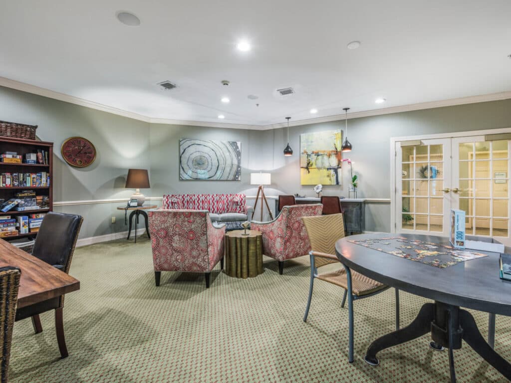 Arbor Terrace of Asheville offers a cozy senior living space with chairs, a sofa, and various tables against light green walls. Artwork adds charm, a game-filled bookshelf invites leisure, and French doors open to another room. The comfortable carpeted flooring enhances this independent living gem.