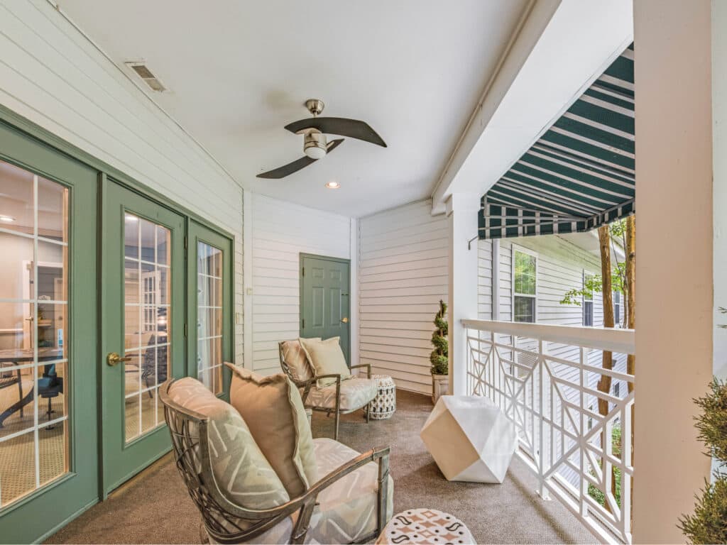 The Arbor Terrace of Asheville features a cozy covered porch in an independent living community, offering cushioned chairs, a small table, and a ceiling fan. Double glass doors with a green awning above a white railing create a relaxing space with modern decor elements perfect for leisurely afternoons.