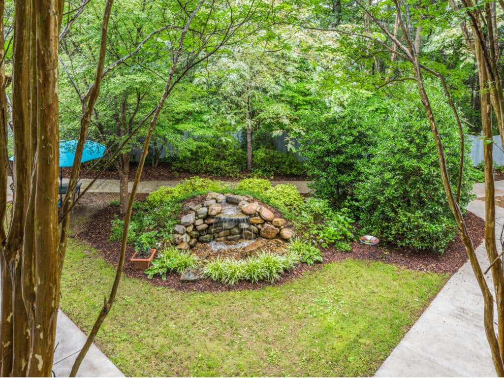 Arbor Terrace of Asheville offers a serene garden with a rock fountain, lush greenery, bushes, and trees. A stone pathway and a blue umbrella over the seating area create an idyllic setting for independent living amidst natures beauty.