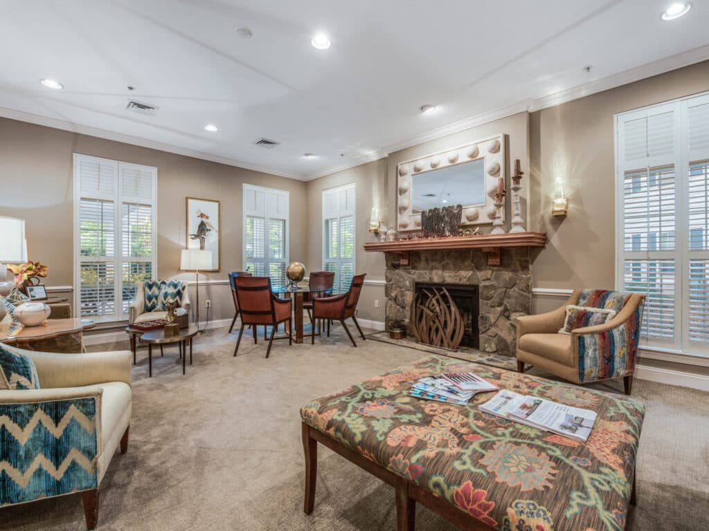 The cozy living room at Arbor Terrace of Asheville features patterned armchairs, a large cushioned bench, and a stone fireplace. A small table with chairs by the windows invites relaxation, complemented by magazines on the bench. Natural light floods the bright space.