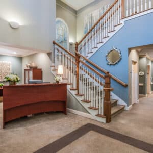 Arbor Terrace of Asheville welcomes guests with a spacious lobby adorned in serene light blue tones. To the left, a wooden reception desk greets arrivals, while a staircase featuring white railings and a wooden banister elegantly ascends. A circular mirror and captivating artwork complete the ambiance.