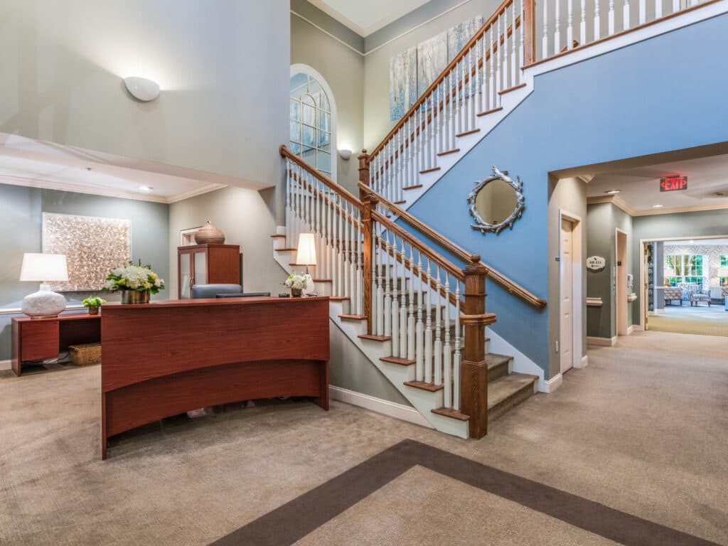 Arbor Terrace of Asheville features a warmly lit office lobby with a wooden reception desk adorned with a lamp and flowers. A light blue staircase and wooden railing lead to an upper level showcasing senior living artwork, while beige carpeting enhances the retirement-friendly atmosphere.