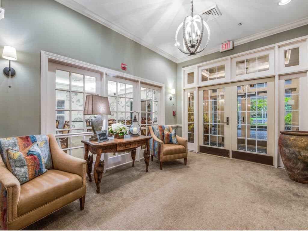 The inviting lobby at Arbor Terrace of Asheville, ideal for independent living in Hendersonville, includes beige armchairs with colorful cushions, a wooden table with a lamp and photo frame, large decorative vases, glass doors with an exit sign above, and a circular chandelier on the ceiling.