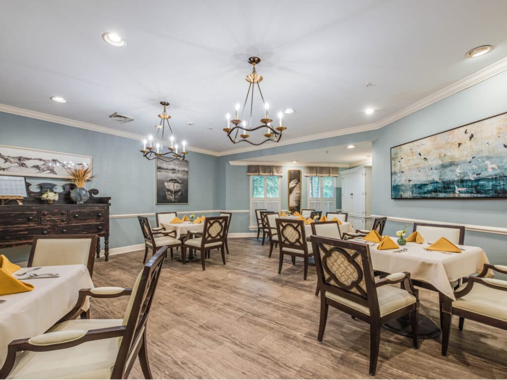 Arbor Terrace of Asheville offers a cozy dining area ideal for a senior living retreat. Wooden floors host four tables with white tablecloths and yellow napkins, surrounded by elegant cushioned chairs. Soft blue walls featuring abstract art and chandeliers illuminate the space beautifully.