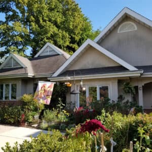The Brooks-Howell is a charming retirement community in Asheville, NC with a gable roof, lush garden, flag, and bird feeder out front. Mature trees frame this serene setting, perfect for those seeking peaceful independent living under clear skies.