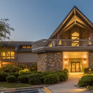 College Walk is a rustic A-frame home in WNC with a stone and wood exterior, warmly illuminated at dusk. A bush-lined pathway leads to the double glass door entrance, while the balcony above features umbrella-topped tables against a clear sky, enhancing the serene ambiance.