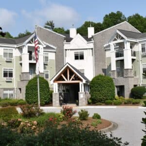 Trinity View in Arden, NC is set amidst greenery, featuring a multi-story structure with green and gray siding. The entrance is highlighted by a peaked wooden canopy, complemented by an American flag nearby, all under a partly cloudy sky for a picturesque setting.