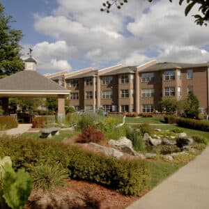 Fletcher Park Inn, a three-story brick building in NC, features many windows and is nestled behind a landscaped garden with pathways, a small pond, rocks, and bushes. To the left, theres a charming gazebo under partly cloudy skies.