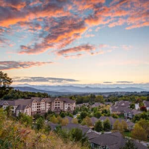 Givens Estates in Hendersonville features residential buildings surrounded by lush greenery. With dramatic sunsets painting the sky in orange and pink and distant mountains on the horizon, it offers an inviting setting for serene senior living.