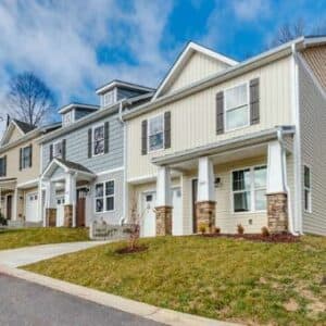 Located in Candler, NC, Liberty Oaks offers modern townhouses with beige and blue exteriors. Each two-story unit includes a small porch with stone accents, a driveway, and is surrounded by lush trees under partly cloudy skies.