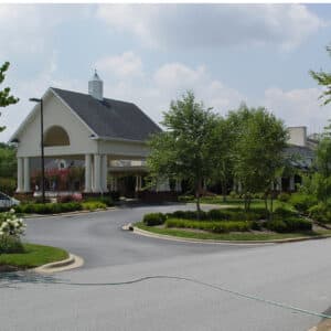 Nestled among trees and lush landscaping, Lake Pointe Landing - Assisted Services boasts a grand building with a covered driveway, white columns, and under a blue sky. A hose spans the driveway beside a parked white vehicle on the left.