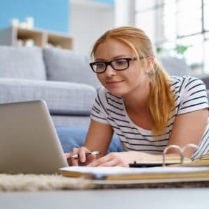 A woman with glasses and a ponytail is lying on the floor, working on a laptop. Shes wearing a striped shirt and jeans, surrounded by notebooks and papers. A gray couch and blue wall serve as the backdrop at Virginia Commons in Arden, NC.
