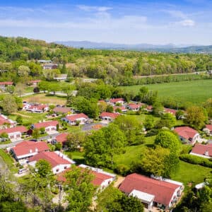 An aerial view reveals Pisgah Valley, a suburban neighborhood with red-roofed houses amid lush greenery. Framed by forests and fields, distant hills under a partly cloudy sky create a picturesque scene reminiscent of NC retirement communities.