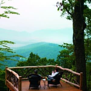 Two people sit on a wooden deck with rustic railings, enjoying the scenic view of lush green mountains and distant mist. Seated comfortably in Southcliff wicker chairs amidst trees, they experience a tranquil atmosphere that connects them serenely with nature.