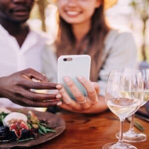 At The Oaks in Arden, NC, a couple enjoys a sunny day at a wooden table, sharing a smartphone. Smiling in the bright outdoor setting, they savor gourmet food and glasses of white wine.