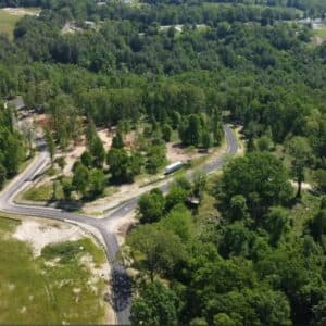 An aerial view of The Orchards at Owensby Road in Hendersonville, NC, showcases a tranquil and verdant environment with winding paved roads meandering through dense woods and open grasslands. Scattered trees enhance the serene and mostly undeveloped landscape.