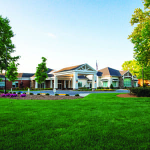 Tryon Estates, a spacious building with a white facade and peaked roof, sits among well-maintained lawns in NC. Surrounded by trees and shrubs under a blue sky, it perfectly captures the essence of retirement living, with a flag waving proudly at its entrance.