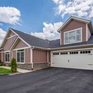 Vistas of Westfield in Candler, NC, features a modern suburban home with a two-car garage under scattered clouds. It combines brick and siding, large windows, and a paved driveway, all surrounded by lush grass and shrubs.