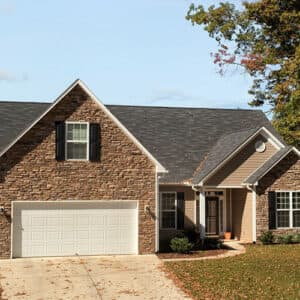 Surrounded by trees, this Fletcher, NC suburban home has a gray roof and stone facade. It boasts a quaint charm with a white-doored two-car garage, three front windows, and a small porch. The grass-lined driveway with scattered leaves is perfect for quiet evenings with The Authors.