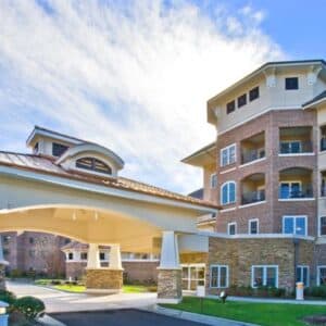 Givens Estates - Assisted Living is a multi-story residential building with a brick exterior, balconies, and large windows, ideal for independent living. It offers a welcoming covered driveway entrance with stone columns under a clear, sunny sky, providing an inviting environment for senior residents.