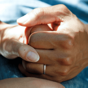 Close-up of two hands holding each other, one with a wedding ring. The softly blurred background captures natural light casting gentle shadows, evoking a peaceful and intimate atmosphere reminiscent of a serene afternoon at Givens Home First in Asheville.