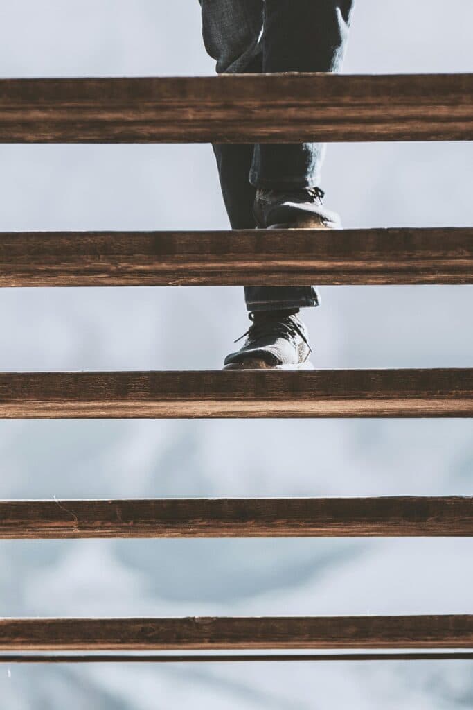 A person wearing dark jeans and shoes is walking down wooden stairs in a serene Hendersonville setting. The perspective is from below, focusing on the lower part of the legs and the stairs against a light background, capturing a moment in this picturesque town.