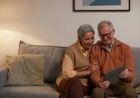 An elderly couple sits comfortably on a sofa in their Asheville home, smiling as they enjoy a tablet together. The softly lit room exudes warmth, with a framed picture on the wall behind them. Dressed in casual yet cozy clothing, they embrace the joys of senior living.