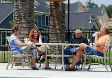 Four people sit around a table under a wooden pergola by the pool at Vistas of Westfield in Asheville. Theyre enjoying sunny chats and sips on outdoor chairs, with stone columns and houses in the backdrop, embracing the relaxed senior living vibe.