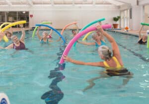 A group of older adults in swimsuits enjoy a lively water aerobics class at Deerfield Episcopal Retirement Community, using colorful foam noodles in the bright and spacious indoor pool, embodying active retirement living in Hendersonville.