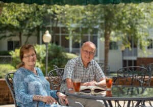 An elderly couple sits at a glass table outdoors at Lake Pointe Landing in Asheville, smiling. Both have drinks; the man reads a book while the woman sits with her hands folded. Trees and a building frame this serene picture of retirement living.