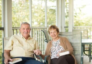 An elderly couple sits on a sunlit porch at Lake Pointe Landing. Holding books and smiling at the camera, they embody retirement joy. Seated on metal chairs, they enjoy a peaceful view of greenery, capturing the serenity of senior living.