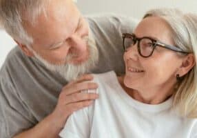 An older couple smiles affectionately at each other, reveling in the joys of retirement in Asheville. The man, with a gray beard, gently rests his hand on the womans shoulder. She has gray hair and wears glasses with her white shirt, set against a softly out-of-focus background.